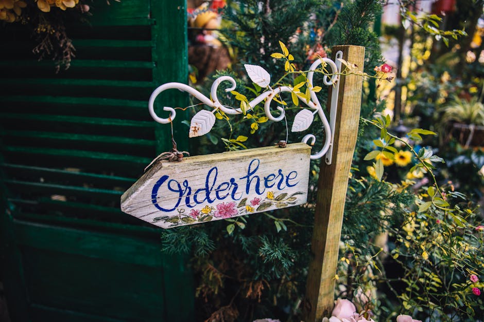 A rustic outdoor garden scene featuring a wooden 'Order here' sign with blue cursive lettering and colorful floral accents hanging from a white wrought iron bracket. Surrounding the sign are lush gree