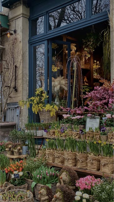 An outdoor flower shop display featuring a variety of fresh flowers and plants arranged on wooden shelves and in woven baskets. The arrangement includes pink, purple, yellow, and white flowers such as