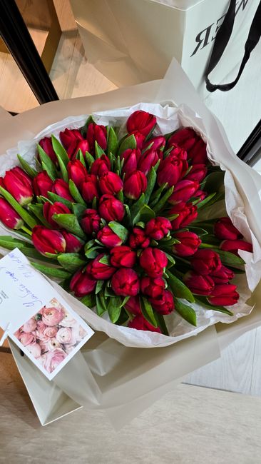A large bouquet of fresh red tulips with vibrant green stems and leaves, arranged in a gathered wrapping of white tissue paper and placed in a white box. The tulips are tightly clustered, showcasing t