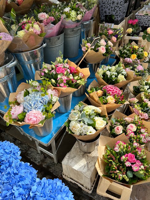 A vibrant display of fresh floral bouquets and arrangements featuring various roses, hydrangeas, and other blooms in soft pinks, whites, and pastel shades, wrapped in brown kraft paper and arranged in