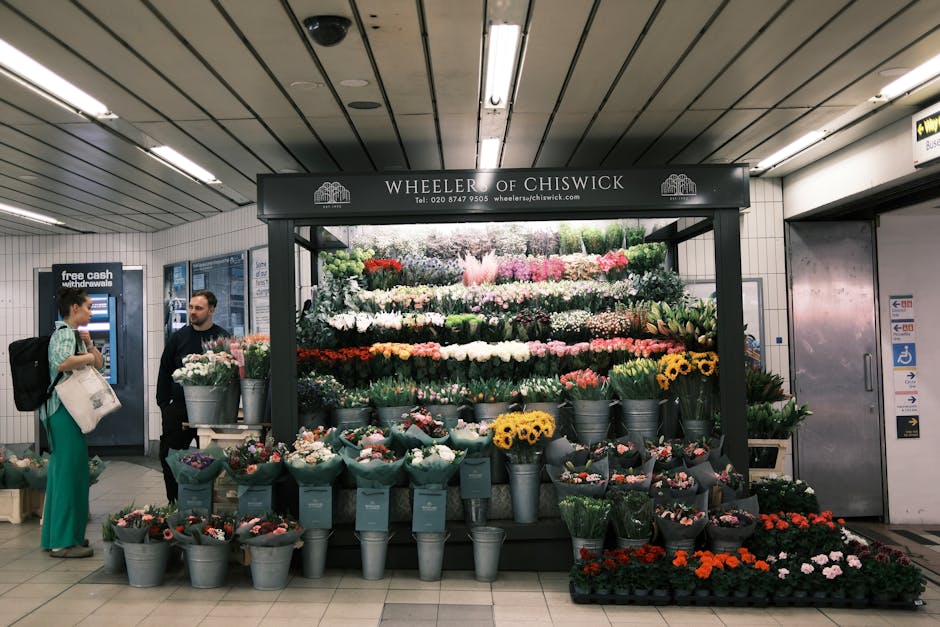 A floral display from Wheeler of Chiswick at Cambridge Heath Station featuring an assortment of fresh flowers arranged in metal buckets and wrapped bouquets. The arrangement includes bright yellow sun