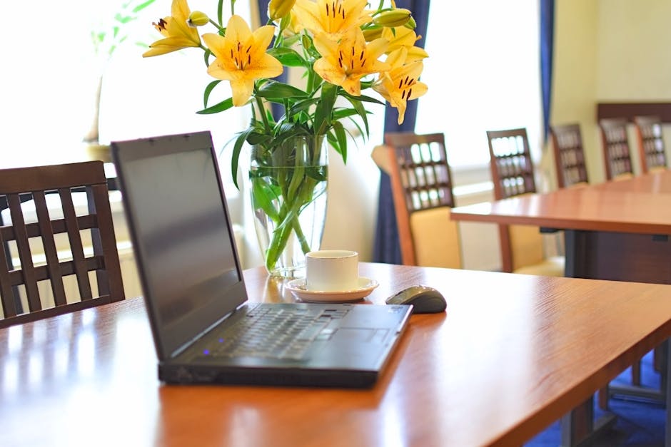 A clear glass vase filled with vibrant yellow lilies and green foliage sits on a polished wooden conference table, surrounded by matching wooden chairs with lattice backs. The arrangement is fresh and